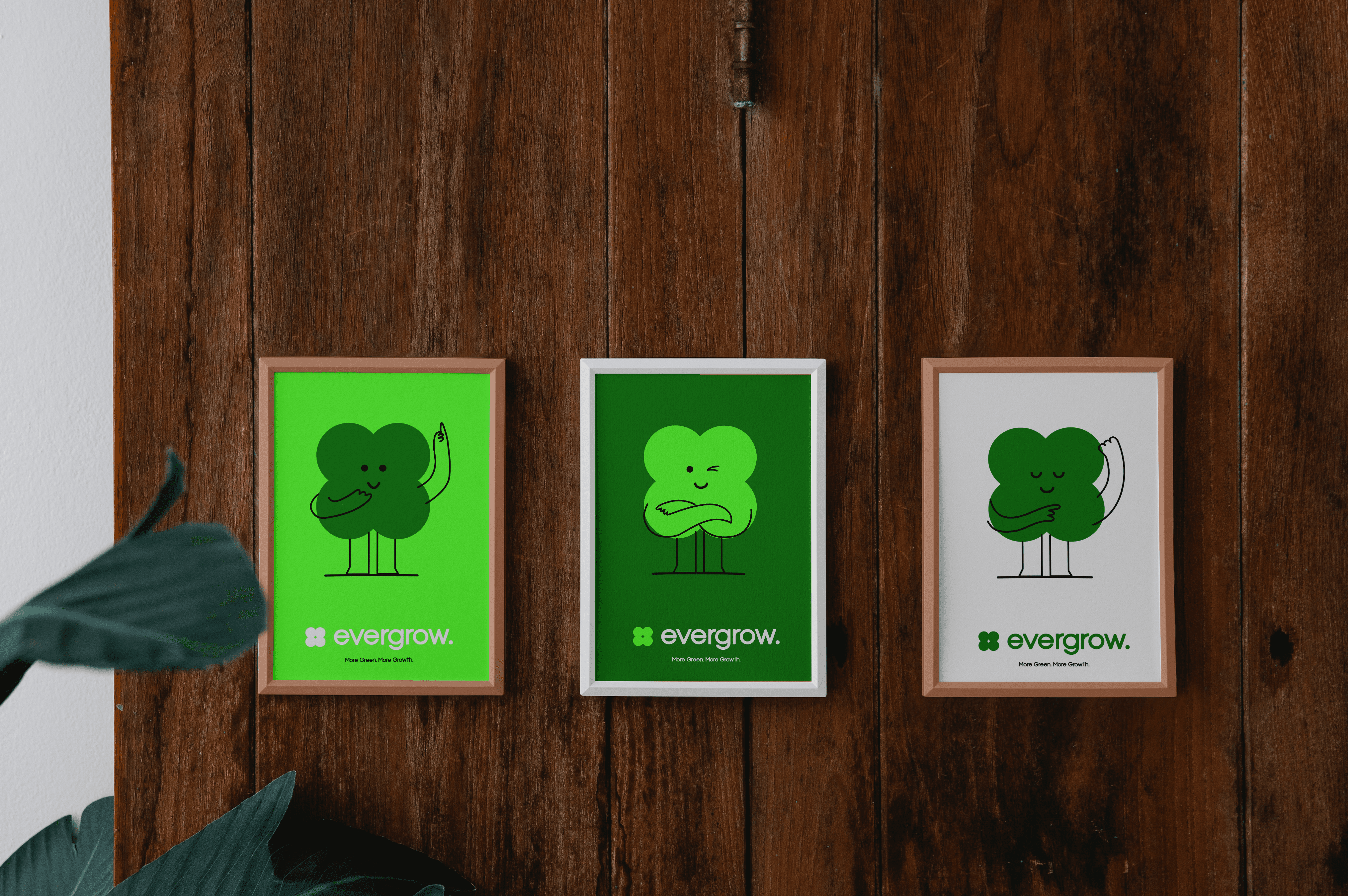 Three framed illustrations featuring whimsical green cloud-like characters with happy expressions are displayed on a wooden wall, with a plant's leaves partially visible at the lower left corner.