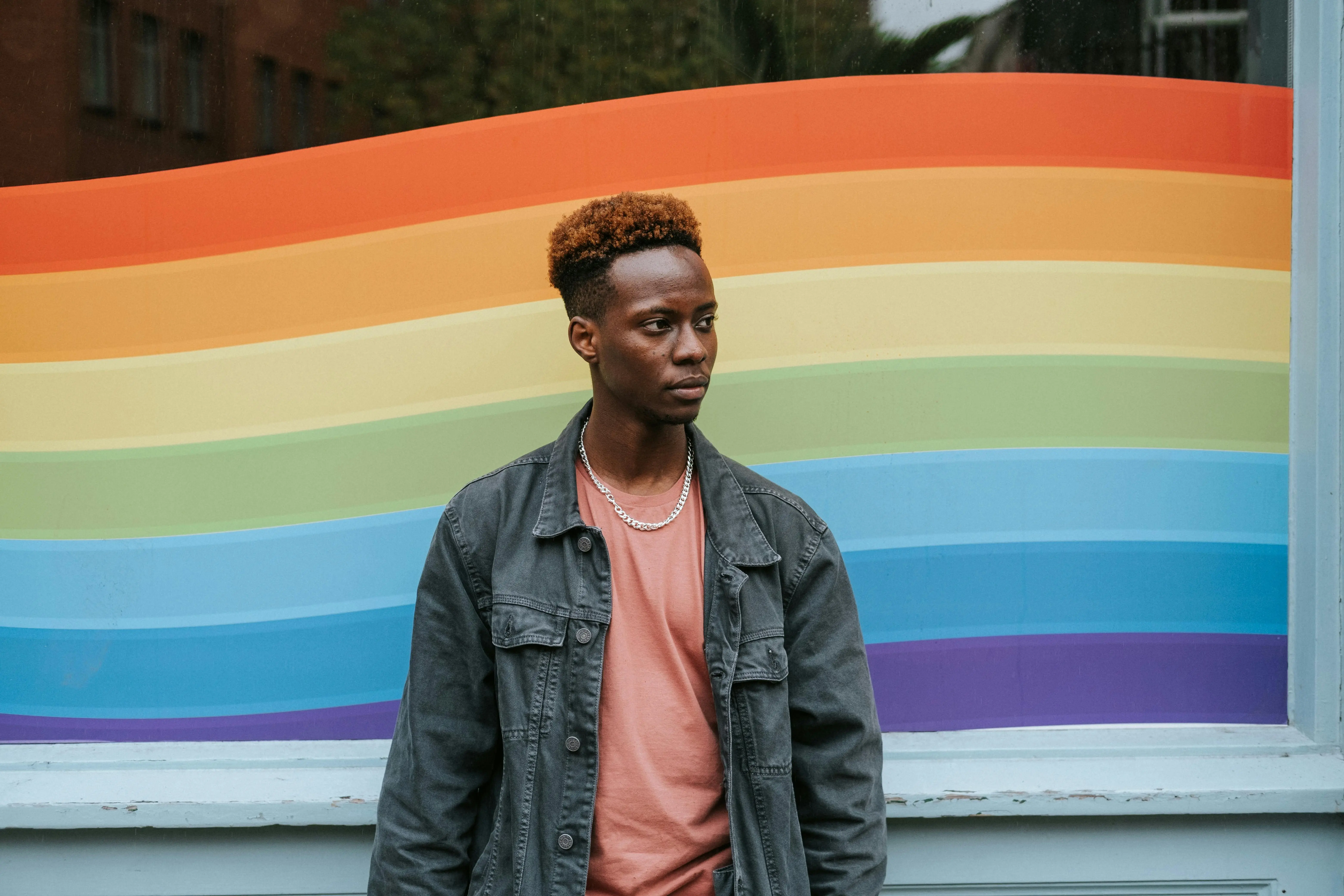 A person stands in front of a vibrant rainbow-colored mural, wearing a denim jacket over a coral shirt, embodying diversity and inclusivity.
