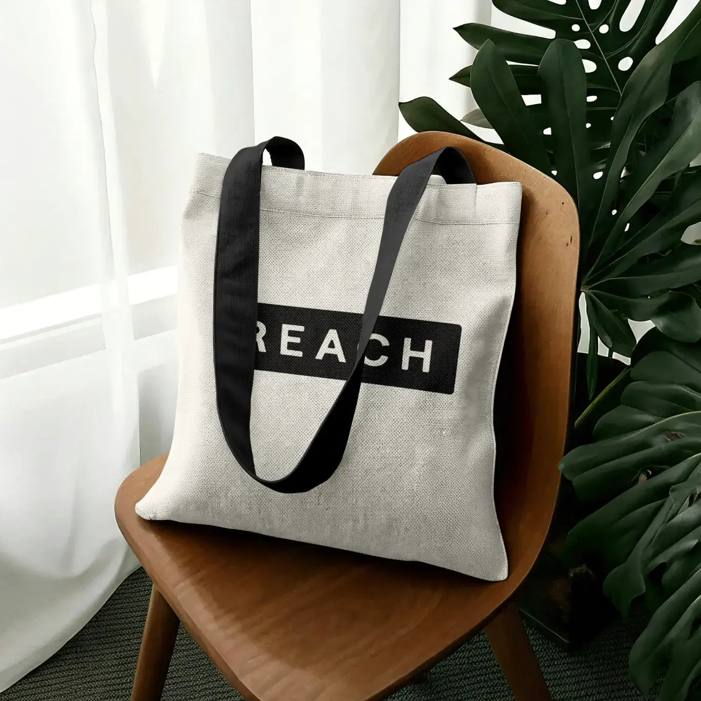 Natural canvas tote with black handles and a bold Reach wordmark, resting on a wooden chair.