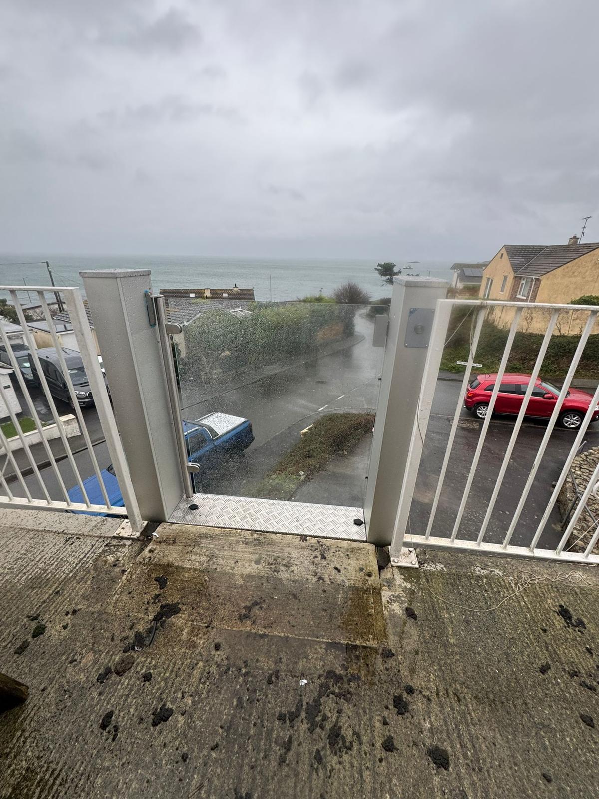 Upper landing view of vertical platform step lift at coastal residential property — glazed enclosure open at balcony level with sea view and residential street below