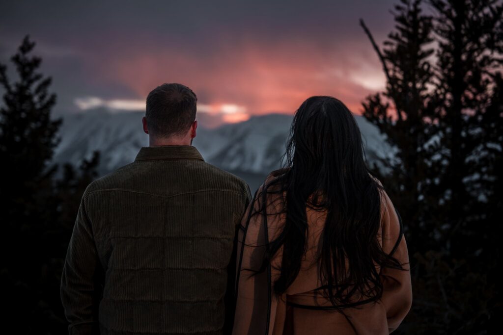 Engaged couple standing with their backs to the camera watching a dramatic red and purple winter sunset over snow-covered mountains above Aspen, Colorado
