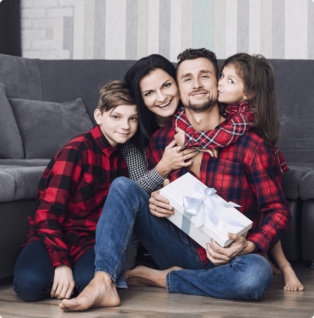 A cheerful family poses outdoors. A father in red holds a child on his shoulders, while a mother in a beige coat cradles a baby. They all smile warmly.