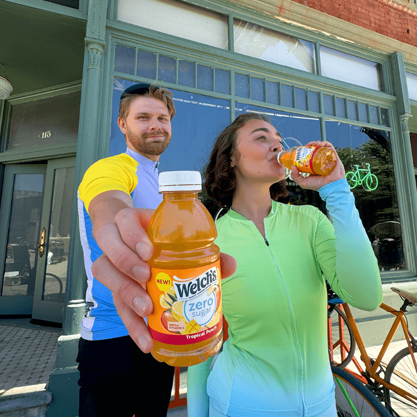 Two people outdoors hold up juice bottles, smiling at the camera, with a bike nearby and a store in the background.