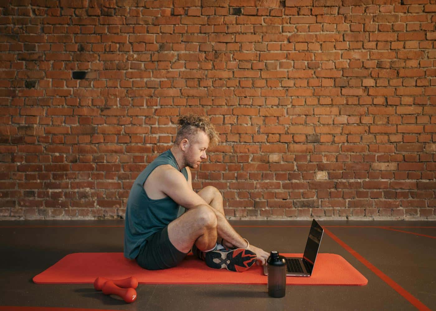 Man sitting on an orange yoga mat with weights next to him looking at open laptop in front of him