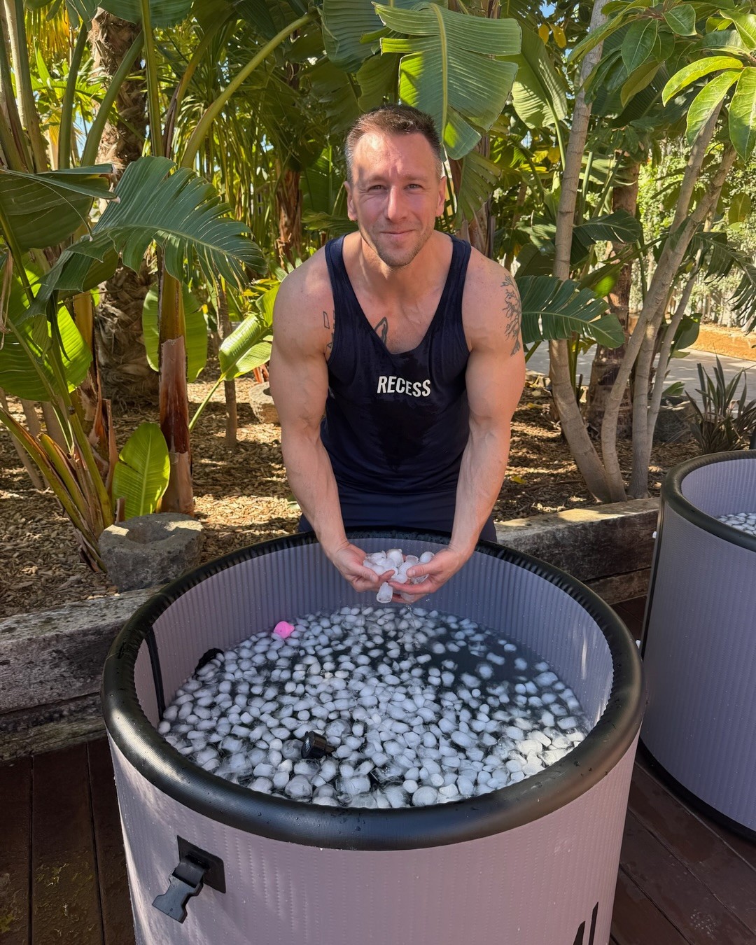 A person stands by an outdoor ice bath filled with ice and wearing a black tank top, surrounded by lush tropical plants under bright sunlight.