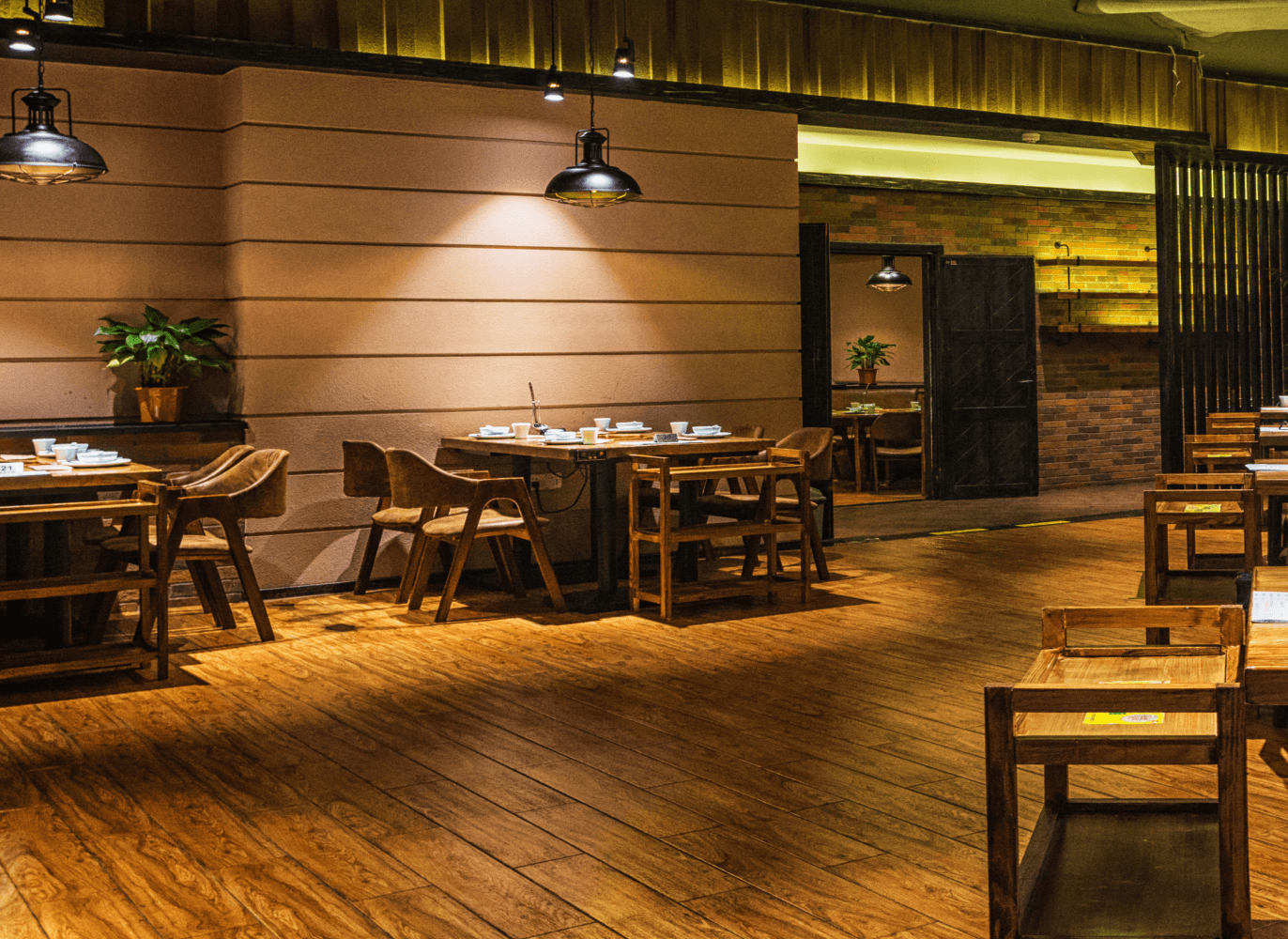 Una vista interior de un restaurante con mesas y sillas de madera, con una iluminación suave. El ambiente se ve acogedor y elegante, con una decoración mínima y una atmósfera cálida.