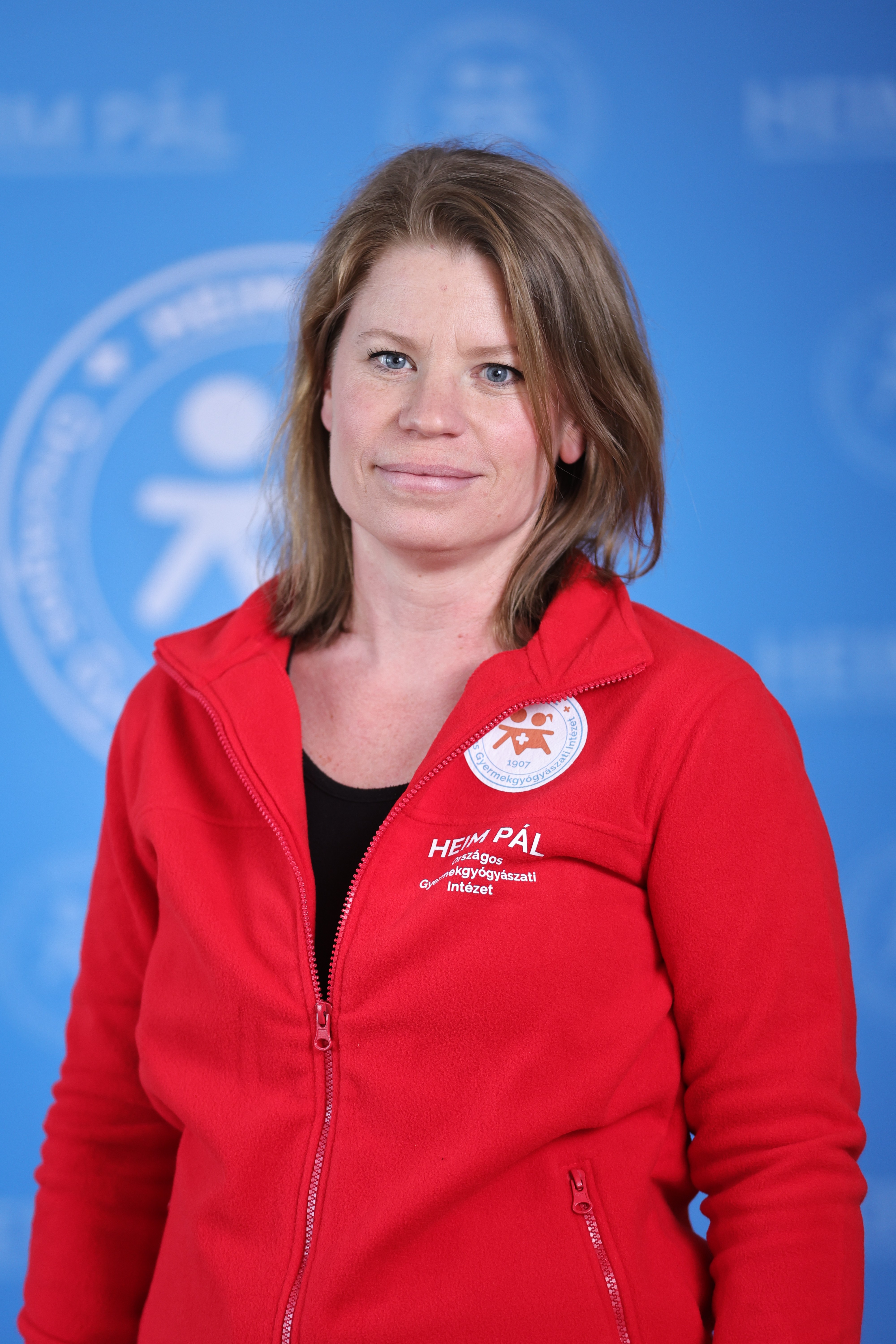 Világos hajú nő mosolyog, piros heim Pál logós pulóverben kék háttér előtt