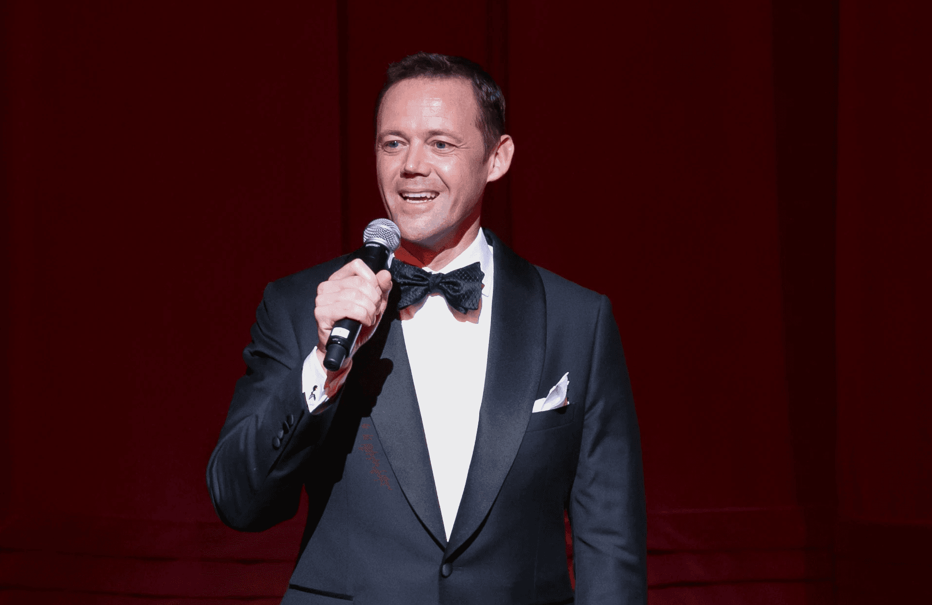 Man in tuxedo in front of opera curtain holding microphone welcoming crowd