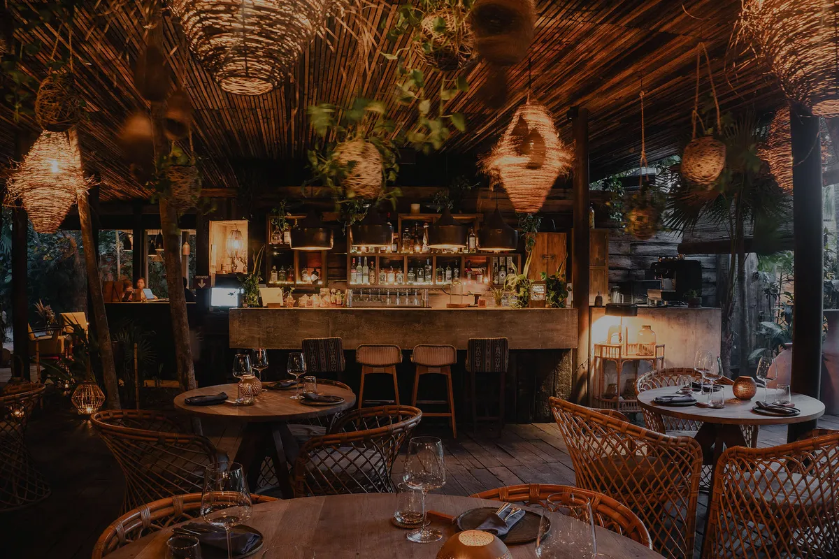 Ocumare restaurant at Be Tulum, serving Mexican cuisine, surrounded by lush greenery and selvatic ambiance, well-lit at dusk with hanging lamps.