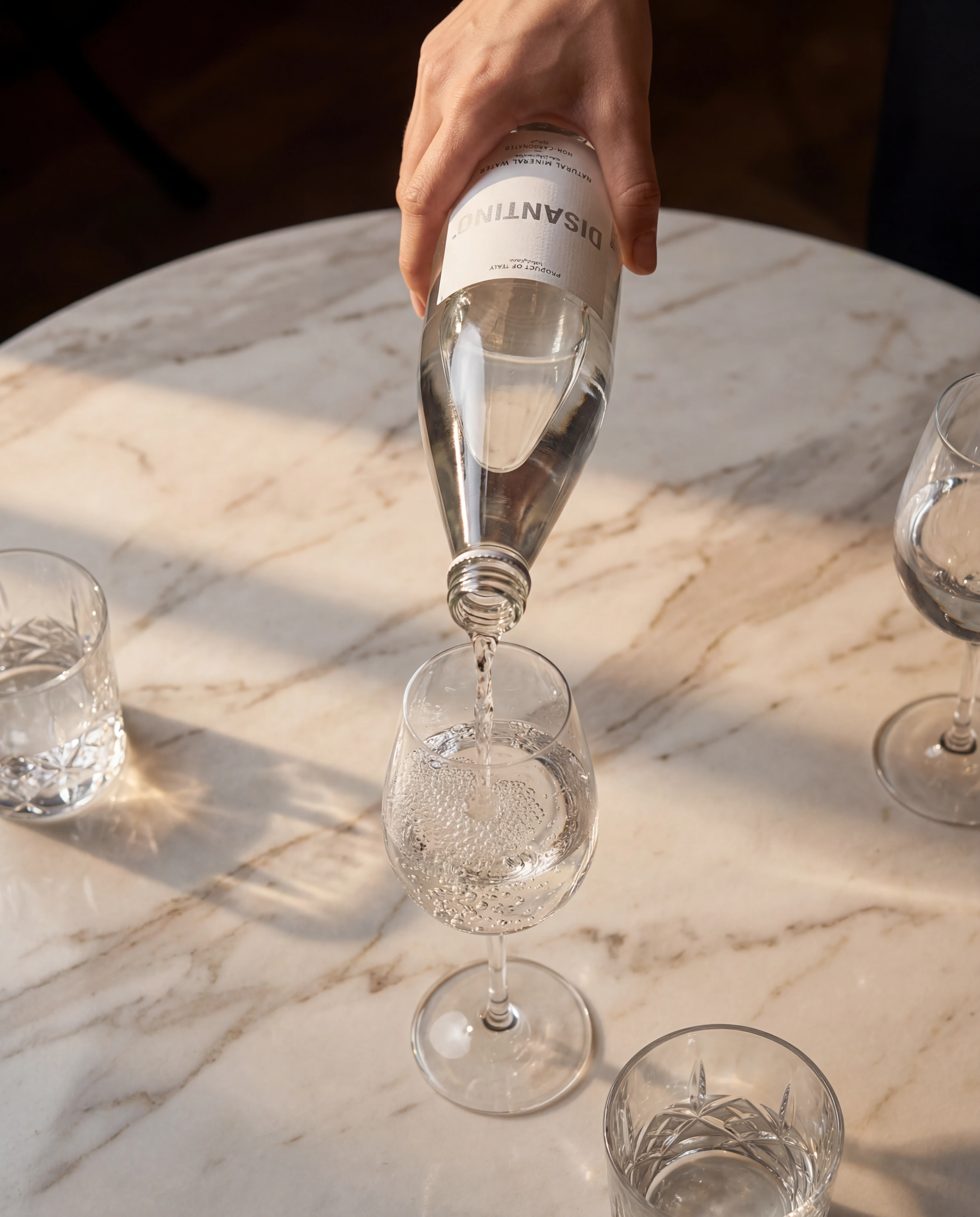 A high-angle view of a hand pouring Disantino water into a stemmed glass on a white marble table. The sunlight casts long, warm shadows across the surface, illuminating the bubbles in the glass and two adjacent crystal tumblers.