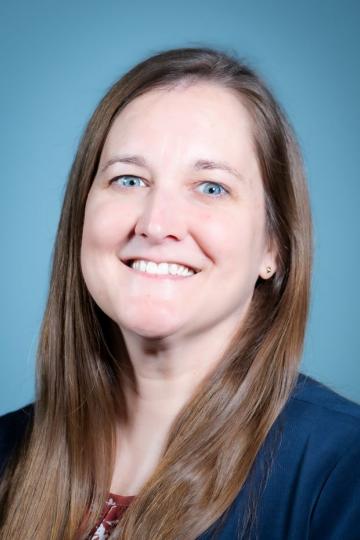 Portrait of a woman with long light brown hair smiling. She wears a navy blue blazer and a floral shirt.