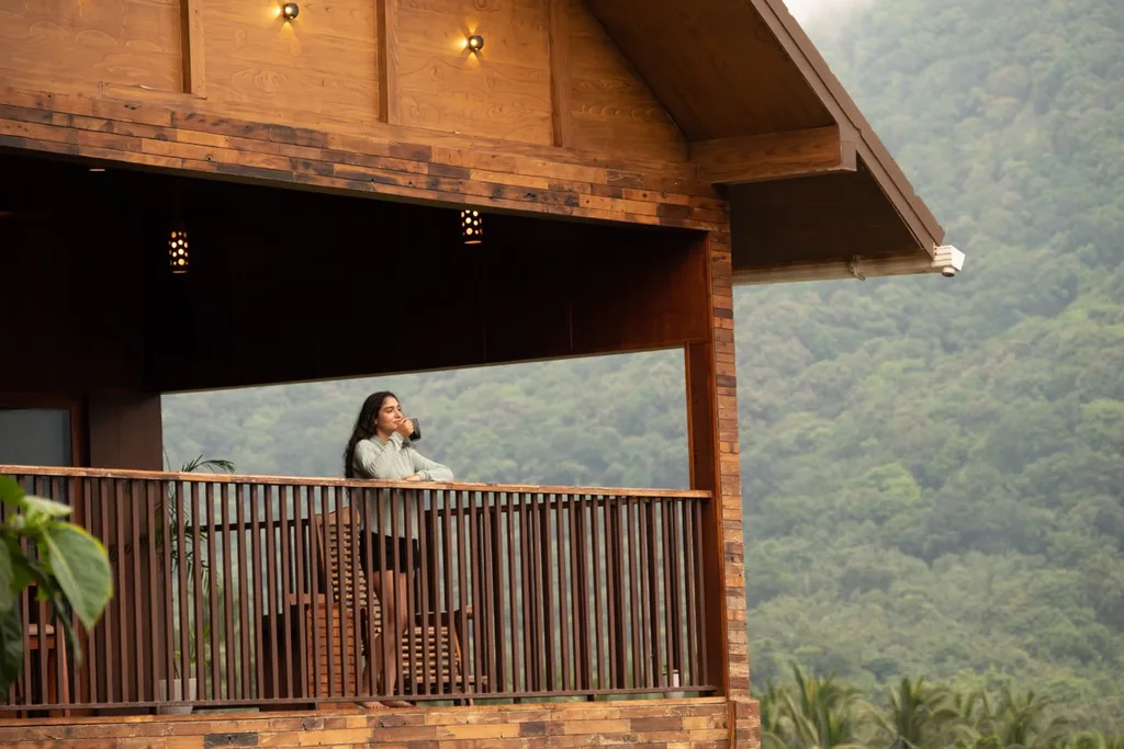 A woman enjoys a cup of coffee while standing on the wooden balcony of the Premium Pool Villa at Au Revoir Wellness Resort, overlooking lush green mountains