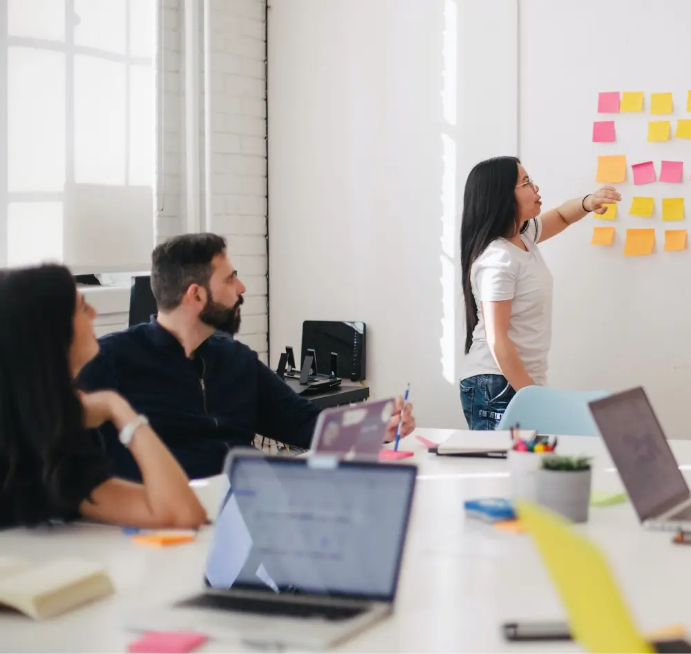 Photograph of woman managing post-it notes on a whiteboard while two other people sit at a desk