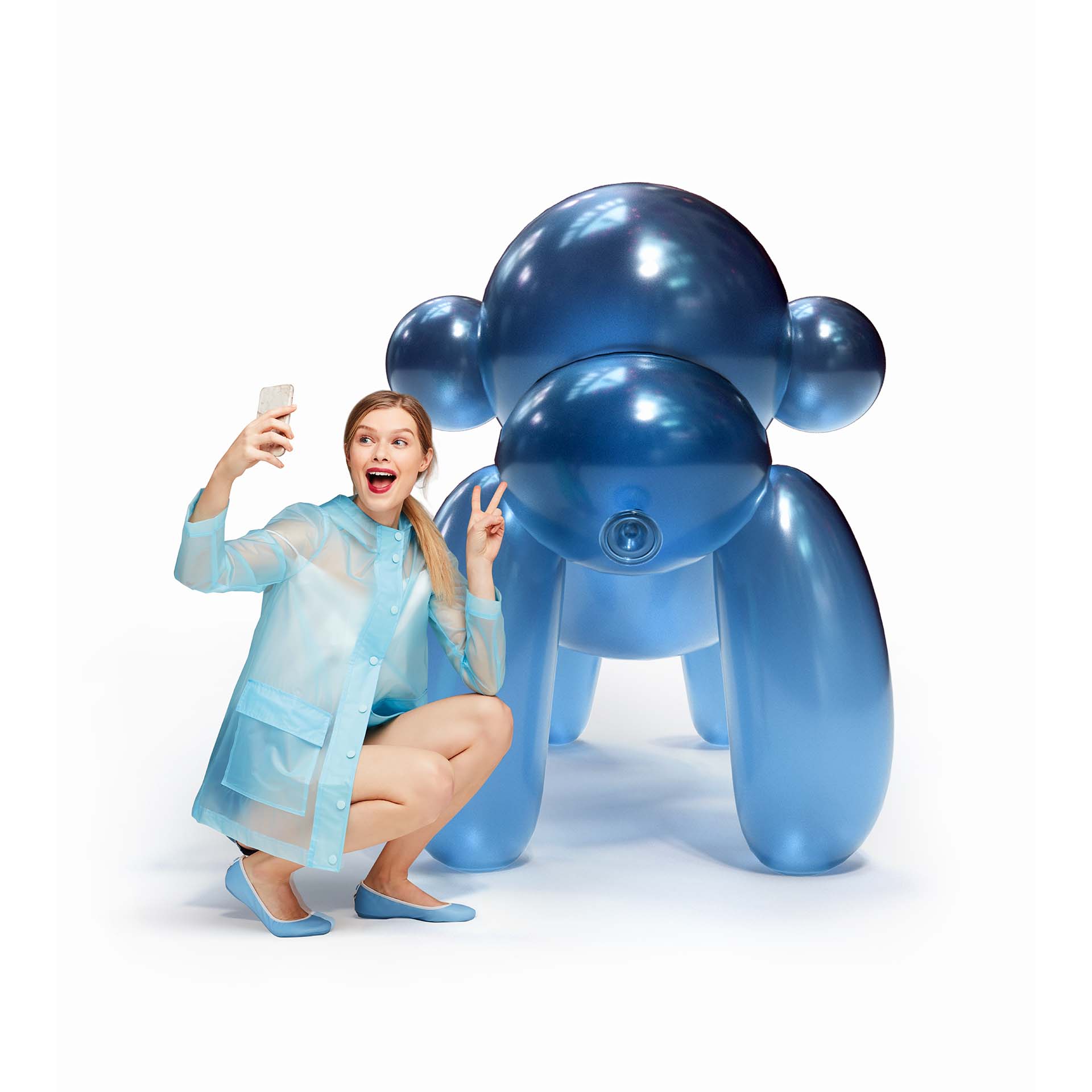 An excited woman in a light blue dress and matching ballet flats takes a selfie with a giant balloon gorilla sculpture in a vibrant metallic blue, flashing a peace sign