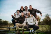 Zes lachende mannen in Broederschap-hoodies op een picknickbank in de natuur