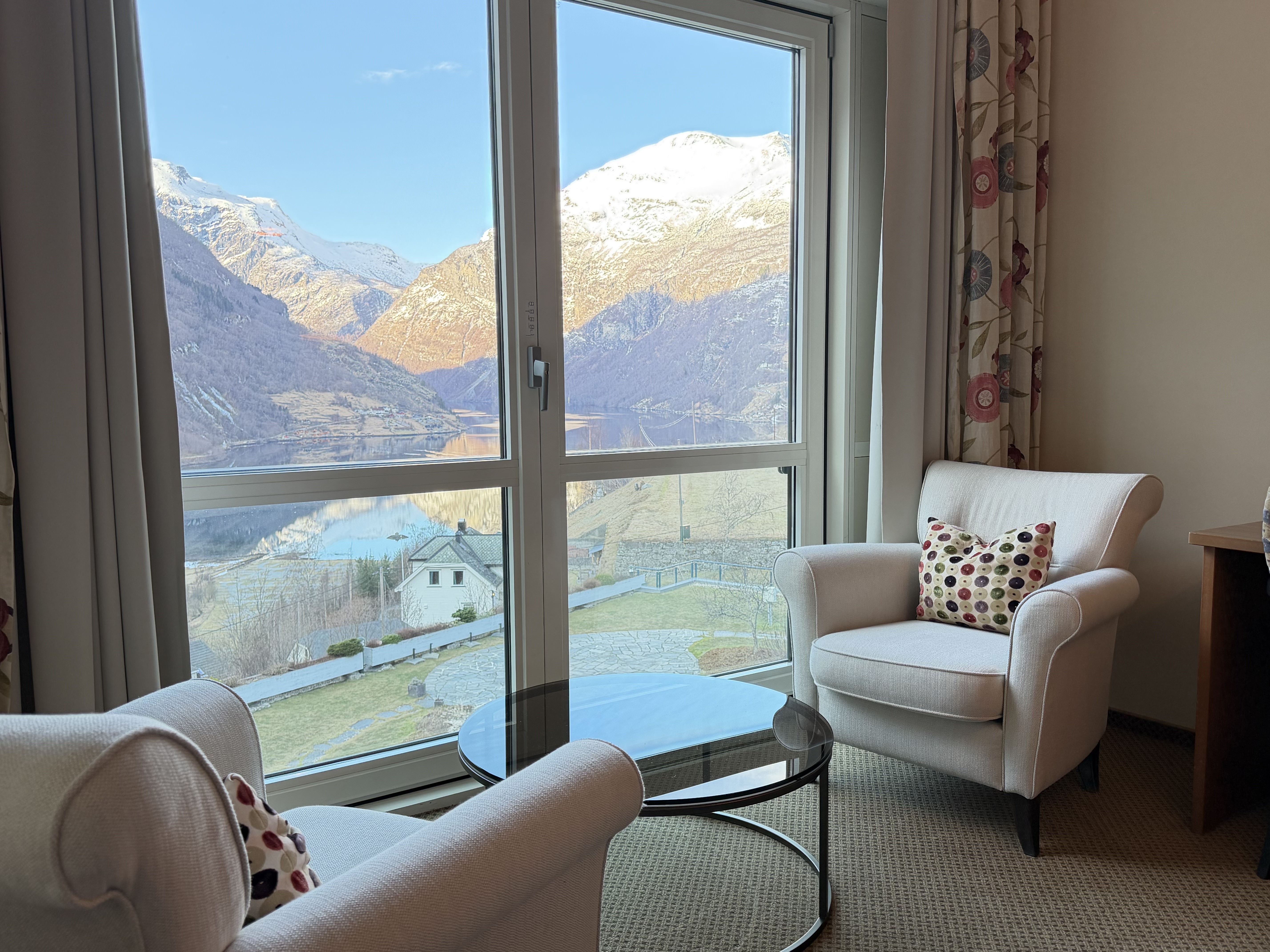 A beautifully designed hotel room features a large double bed with a patterned blue headboard, adorned with decorative pillows, set against a floral accent wall, and complemented by a window view of lush greenery, alongside two plush lounge chairs and a circular table.