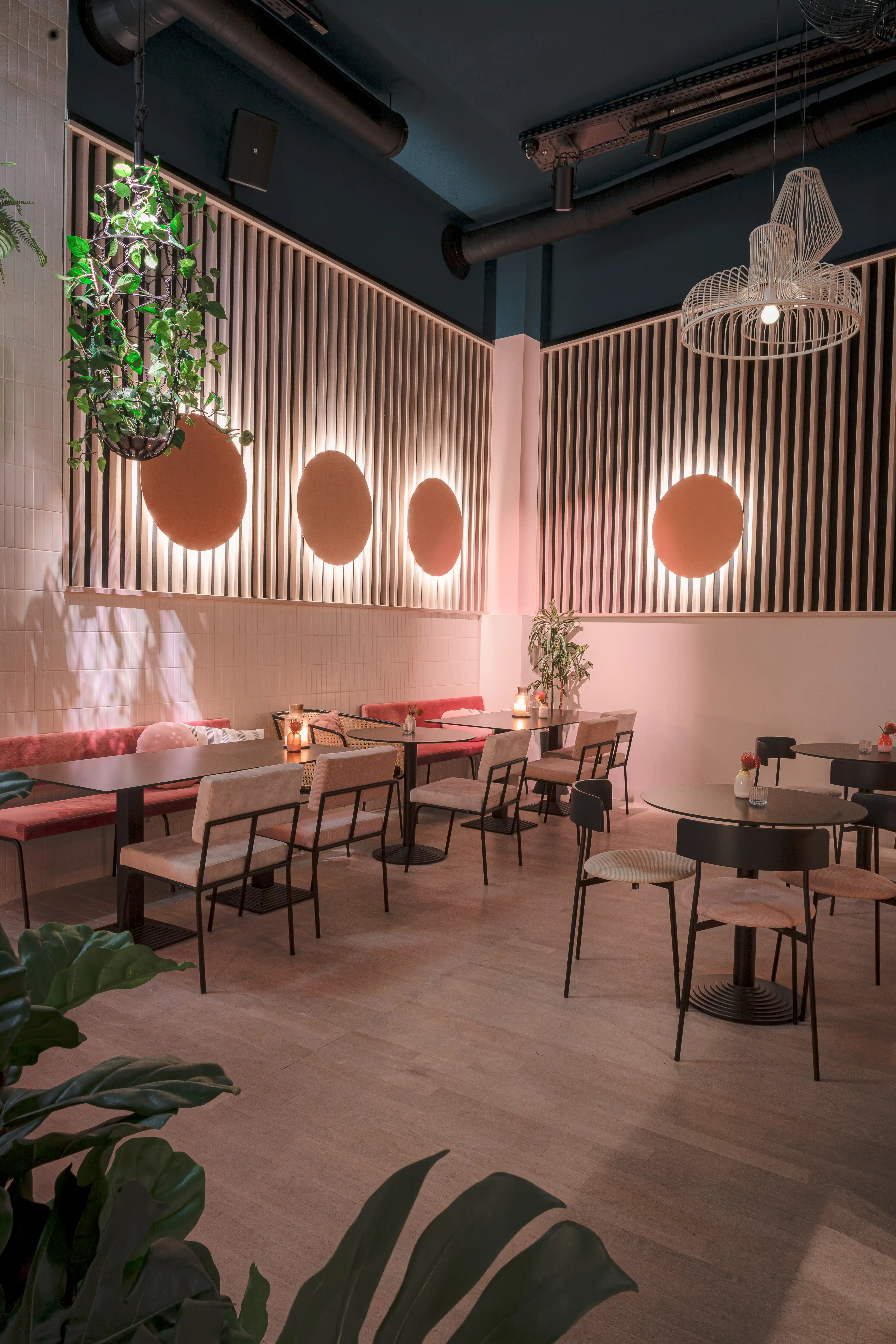 Evening mood at LOKAL VIER with warm-lit copper circles on backlit wooden slat walls, a pink banquette, black bistro tables and a hanging plant under a dark teal ceiling.