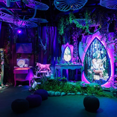 A whimsical, dimly lit room with a fantasy theme, featuring glowing purple decor, plants, and fairy-like imagery on mirrors and screens.