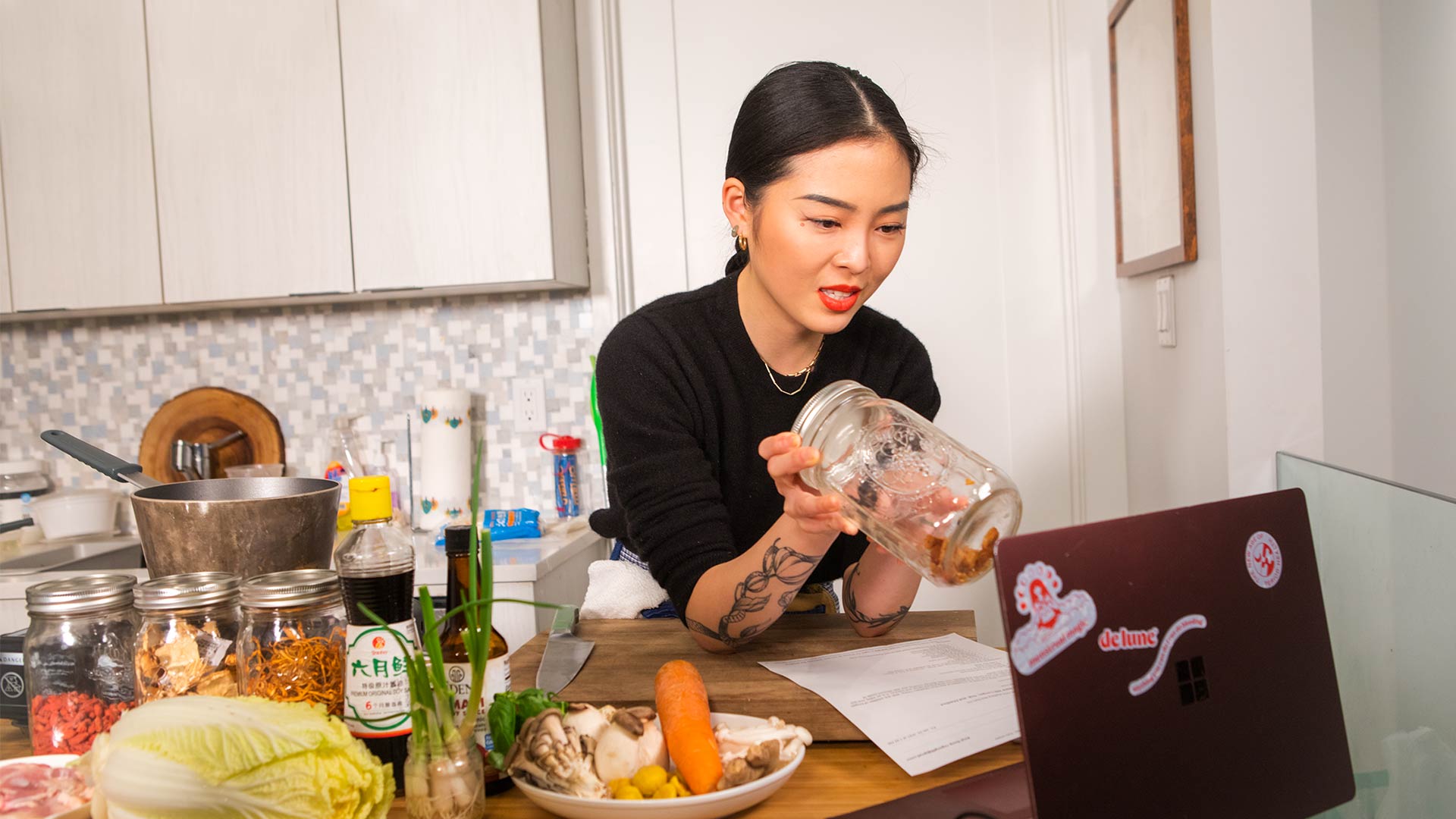 A woman with dark hair pulled back in a ponytail, wearing a black long-sleeved shirt, is pouring a clear liquid from a large glass jar into a smaller glass jar on a light wood countertop. She has red lipstick and a tattoo on her left forearm. Various food items, including vegetables and packaged goods, are visible on the countertop, along with a laptop displaying stickers. A white tile backsplash is behind the counter, and white cabinets are visible above the backsplash.