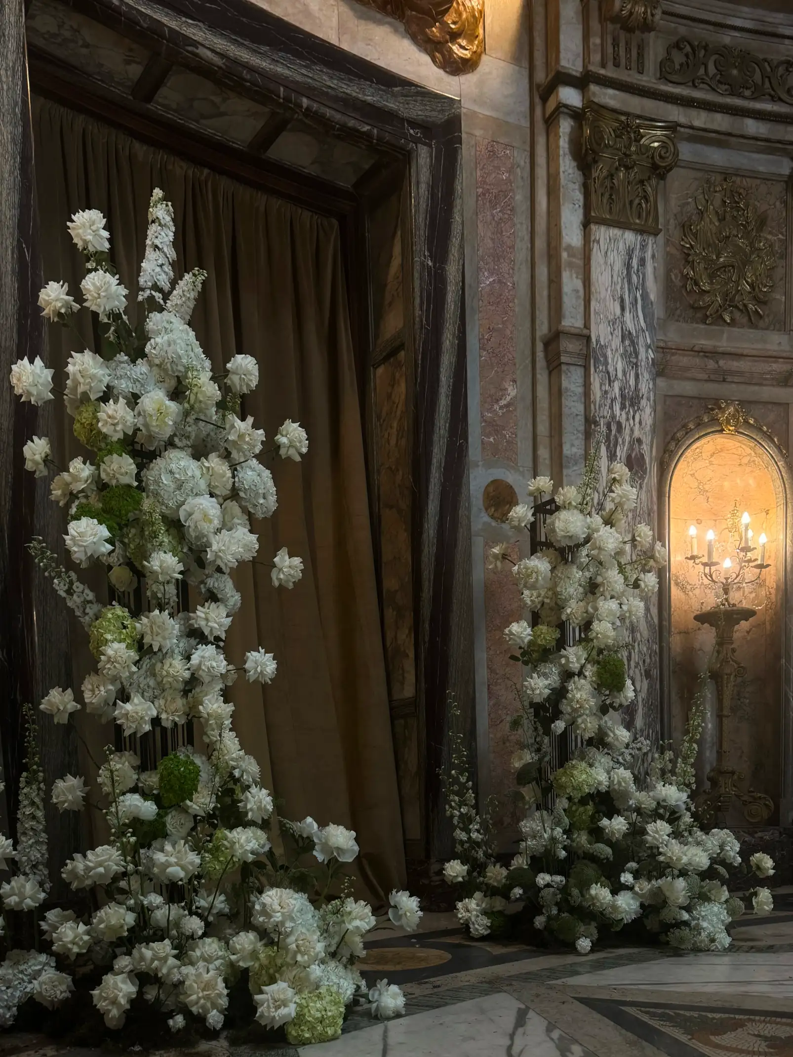 Arreglo floral vertical blanco con flores mixtas y follaje verde en espacio arquitectónico, para ambientación solemne y refinada.