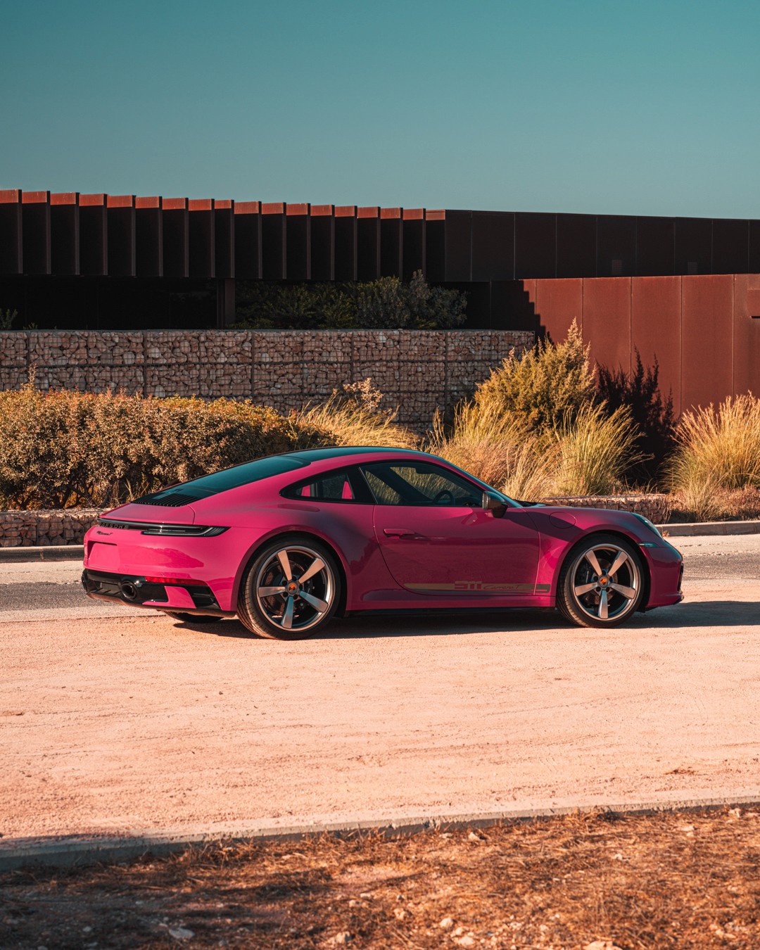 Ruby Star Porsche 911 Carrera T (992) in side profile, rear three-quarter, parked on sandy ground beside ornamental grasses and a corten-steel building, gold "911 Carrera T" side script and Fuchs-style wheels in warm daylight.