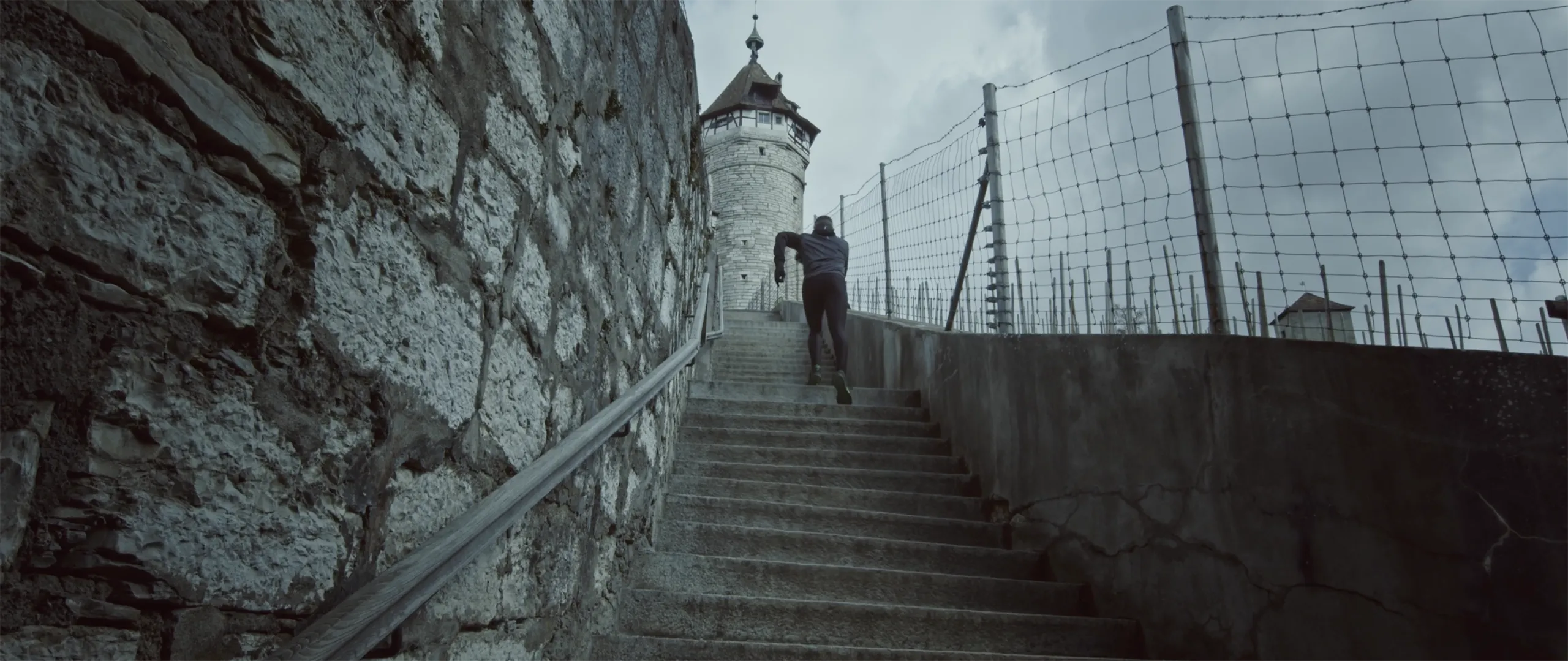 A wide shot of a person running up steep stone stairs next to a historic wall leading to a Munot in Schaffhausen.
