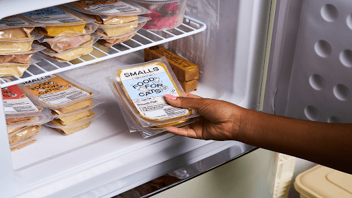 Close-up of a person reaching for a "Small's" brand pre-packaged meal from a refrigerator shelf stocked with similar containers.