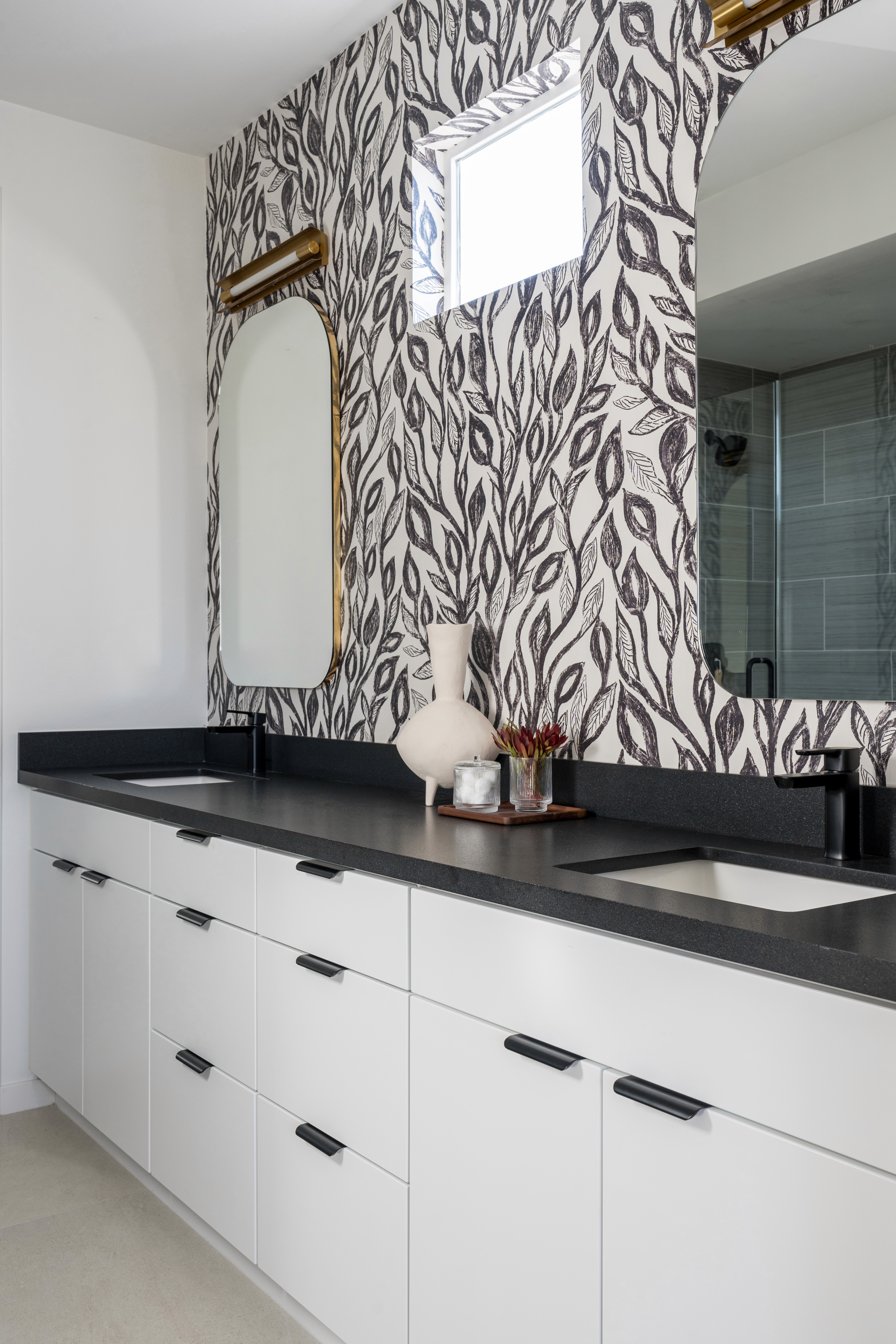 Bathroom vanity with gold framed arched vanity mirror, black and white leaf wall paper, brass sconce lighting, black countertops and flat white cabinets with black hardware