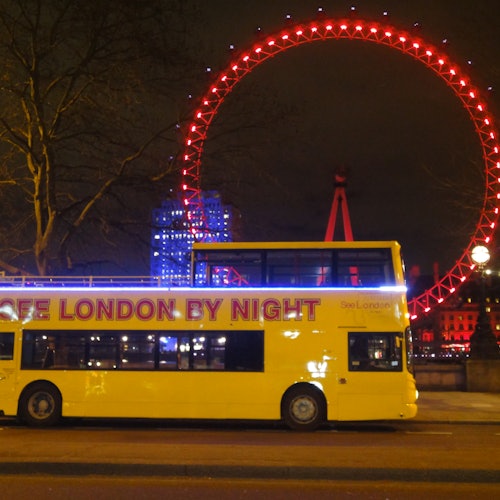 A yellow double-decker bus with "See London by Night" signage, in front of the illuminated London Eye at night.
