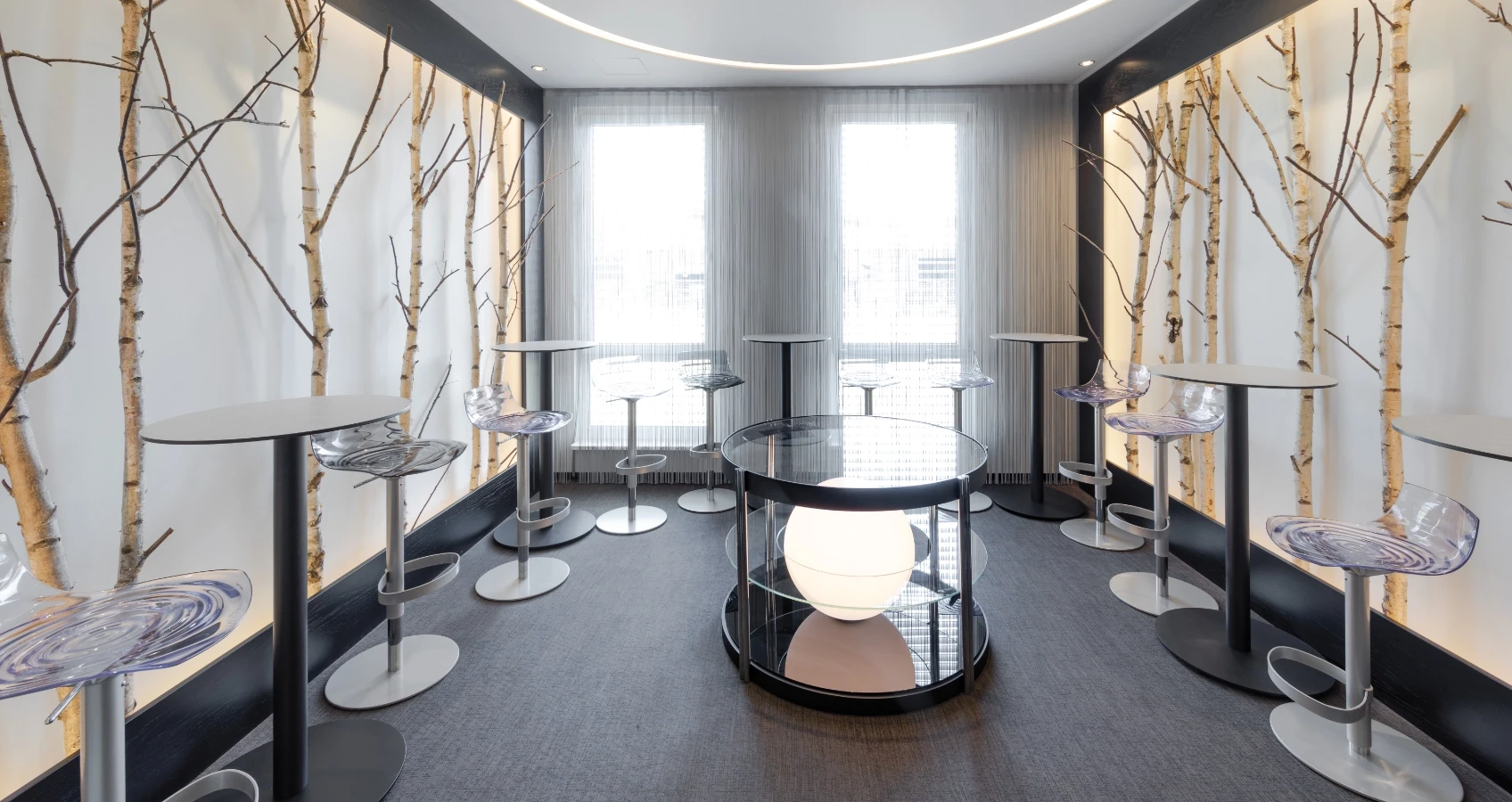 Moderne Lounge mit Birken-Wanddekor, transparenten Barhockern, Stehtischen und runden Glas-Couchtischen mit beleuchteter Kugel.