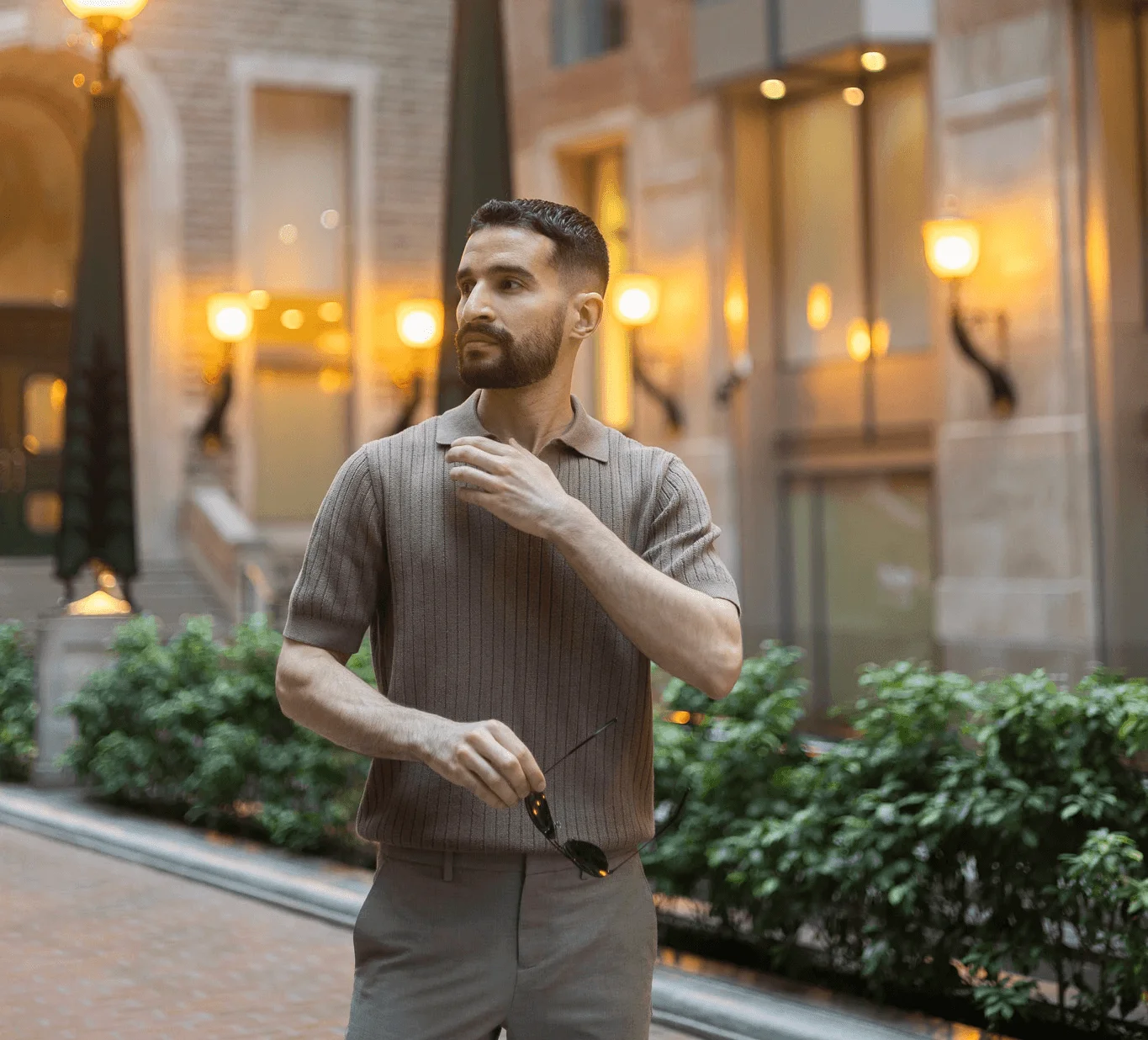 A man in a casual outfit stands outdoors near a building, adjusting his clothing with warm lights in the background.