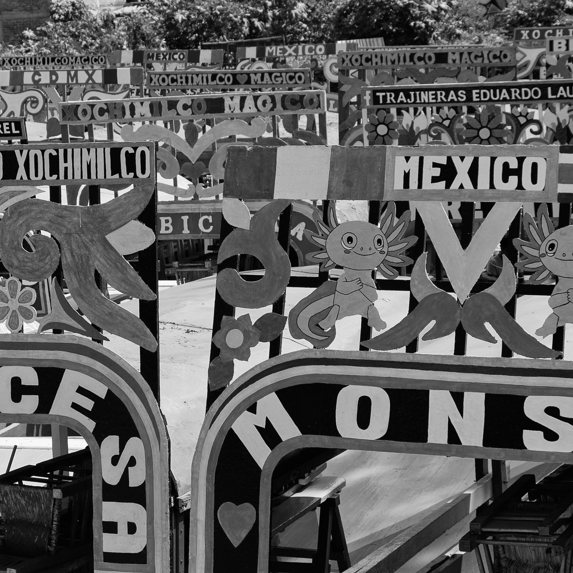 Black and white photo of colorful trajinera arches at Xochimilco, featuring decorative lettering, floral motifs, and axolotl illustrations with the word “MEXICO” prominently displayed.