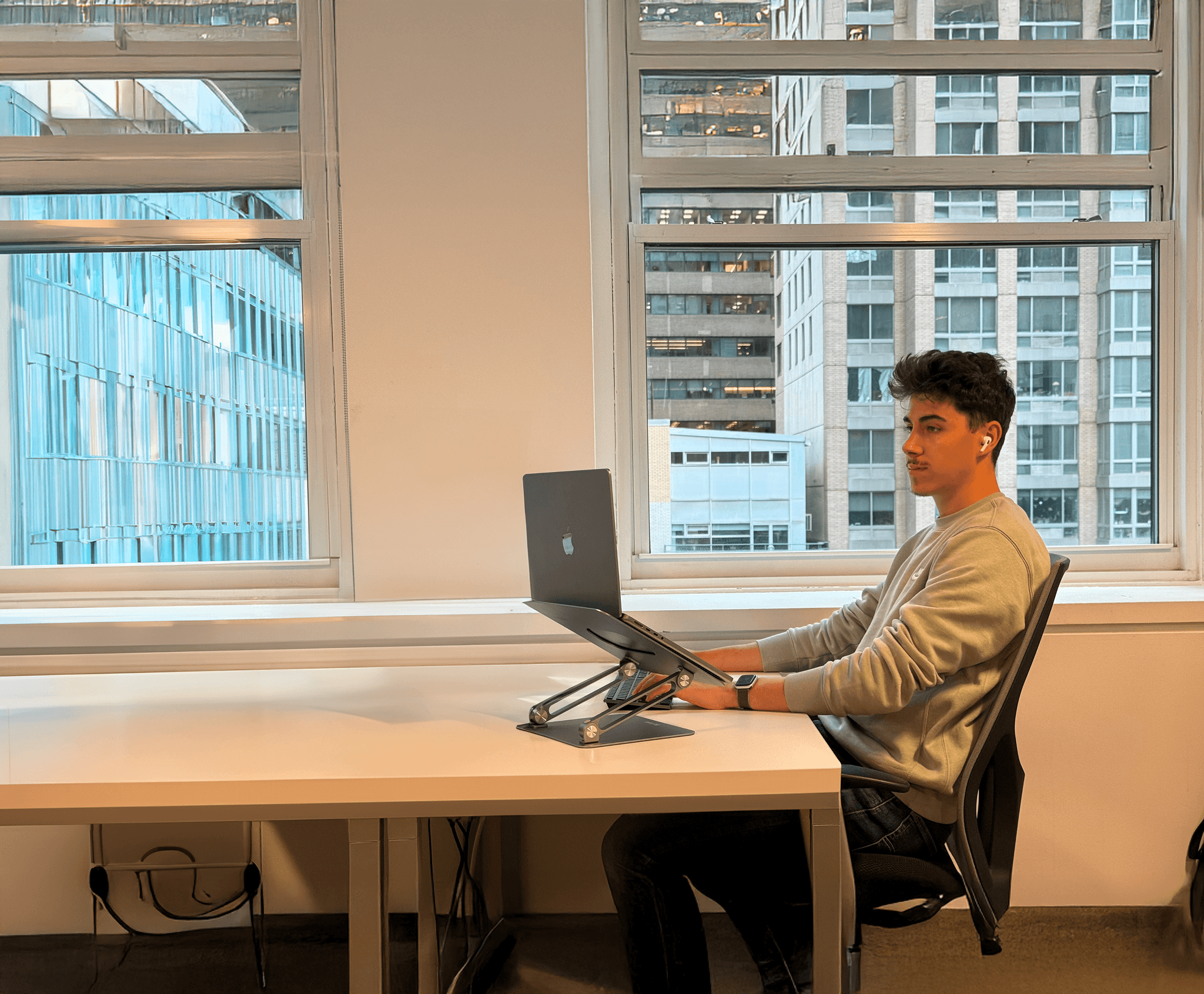 Freelance assis à son bureau dans un open space moderne, travaillant sur un MacBook sur support ergonomique, avec vue sur des immeubles urbains.