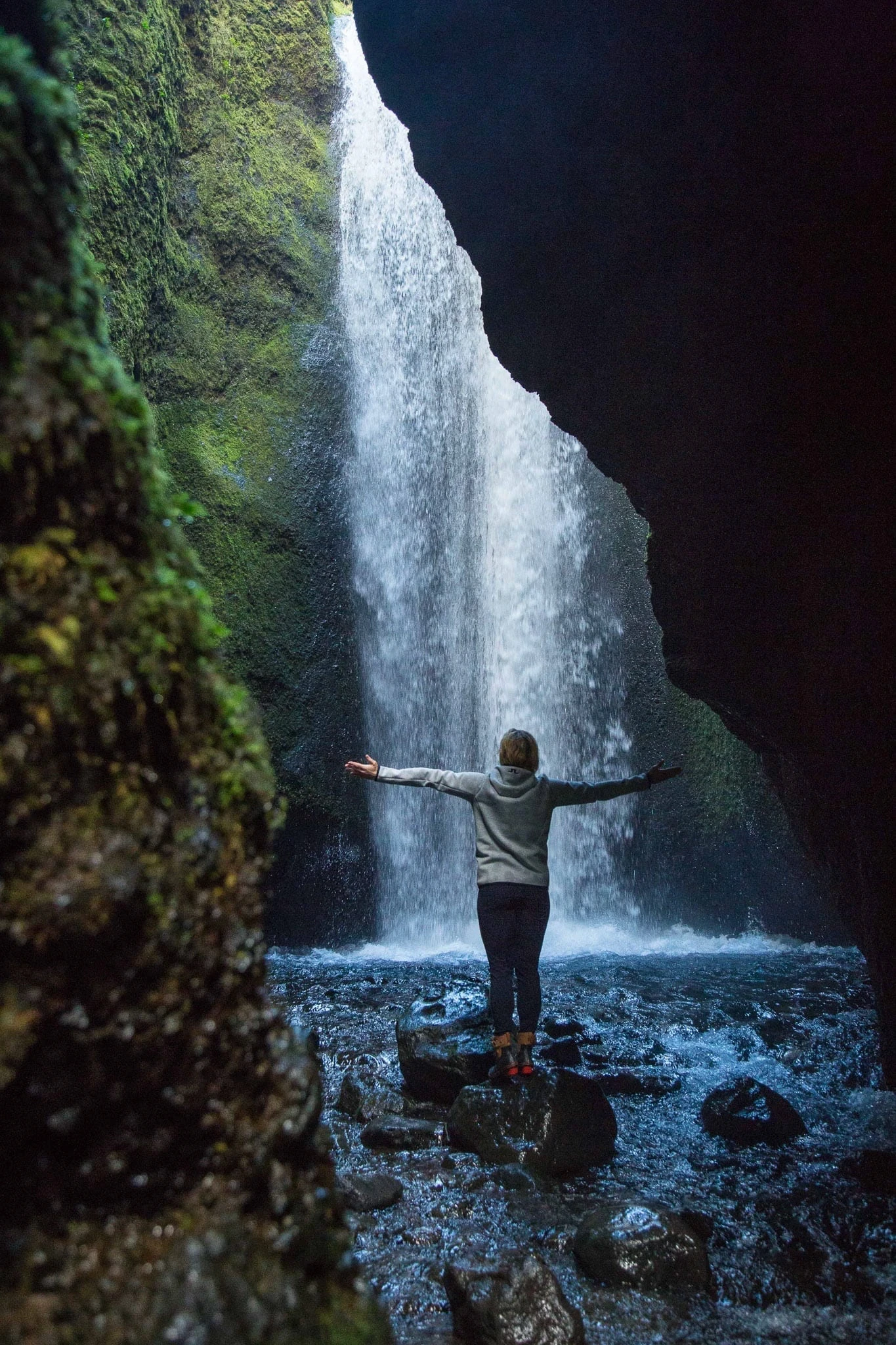 Person standing on a rock with arms outstretched beneath a waterfall.