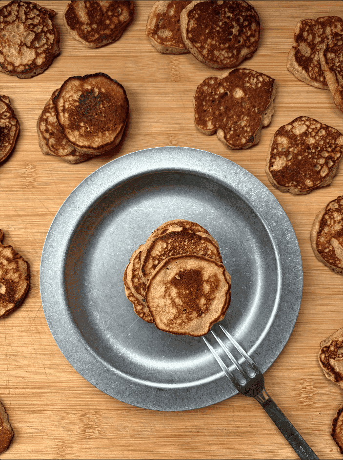 Stack of small golden-brown chickpea pancakes on a metal plate, surrounded by additional mini pancakes on a wooden surface.