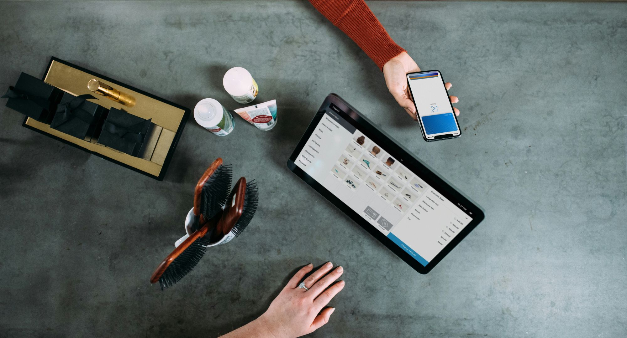 visão de cima de uma mesa com objetos variados e um tablet e uma mão segurando um celular, fazendo um pagamento