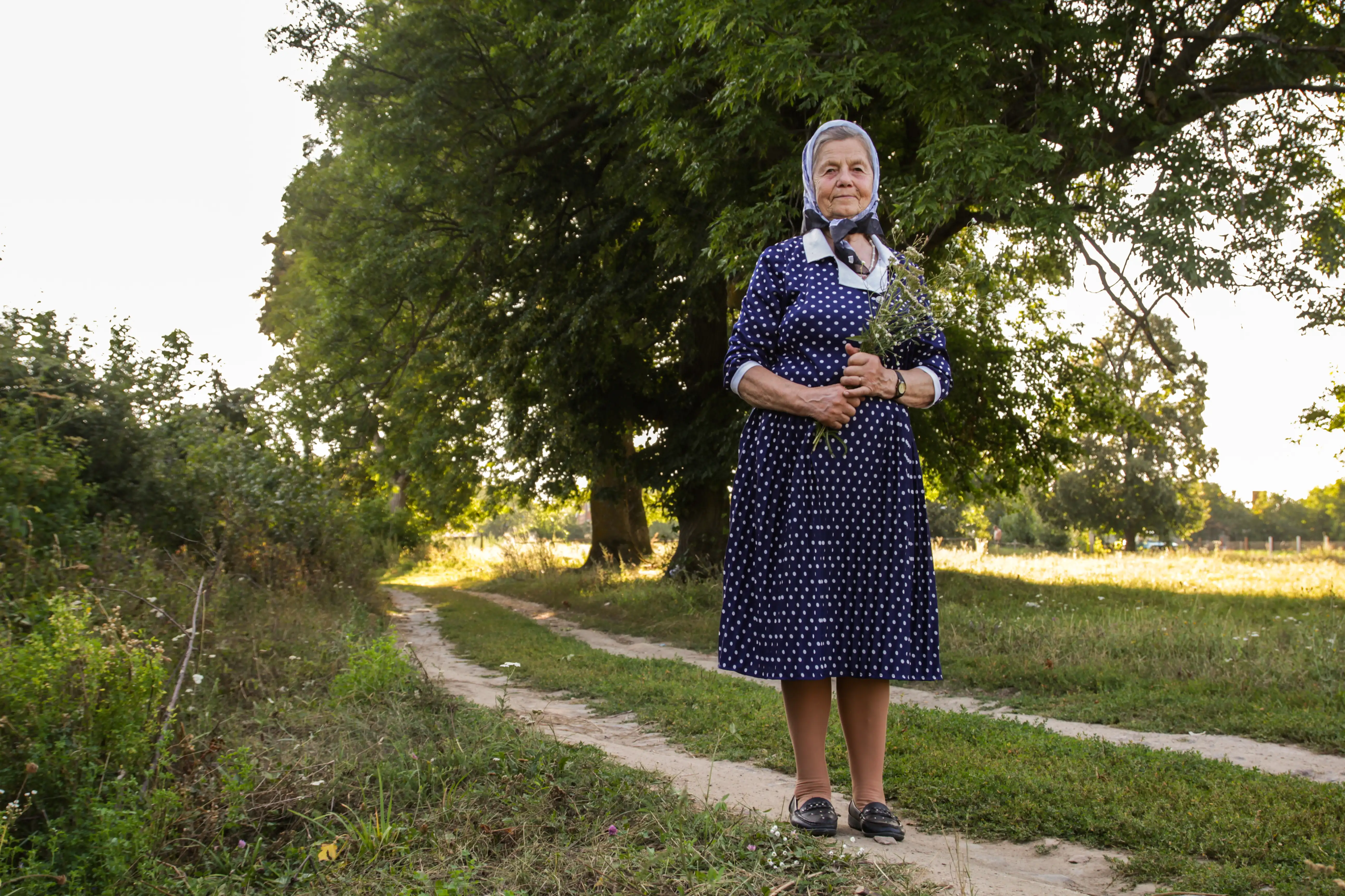 Starsza kobieta w niebieskiej sukience w białe kropki stoi na wiejskiej ścieżce, trzymając kwiaty. W tle widać zielone drzewa i pola.