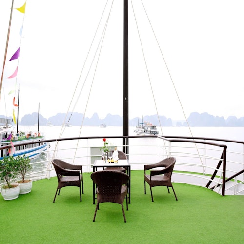 Udendørs terrasse med grøn gulvbelægning, tre brune fletstole rundt om et bord, potteplanter og både på vandet i baggrunden.