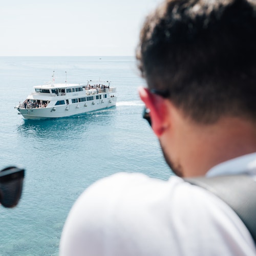 Muž s dalekohledem sleduje bílý trajekt plný pasažérů plující na klidném moři.