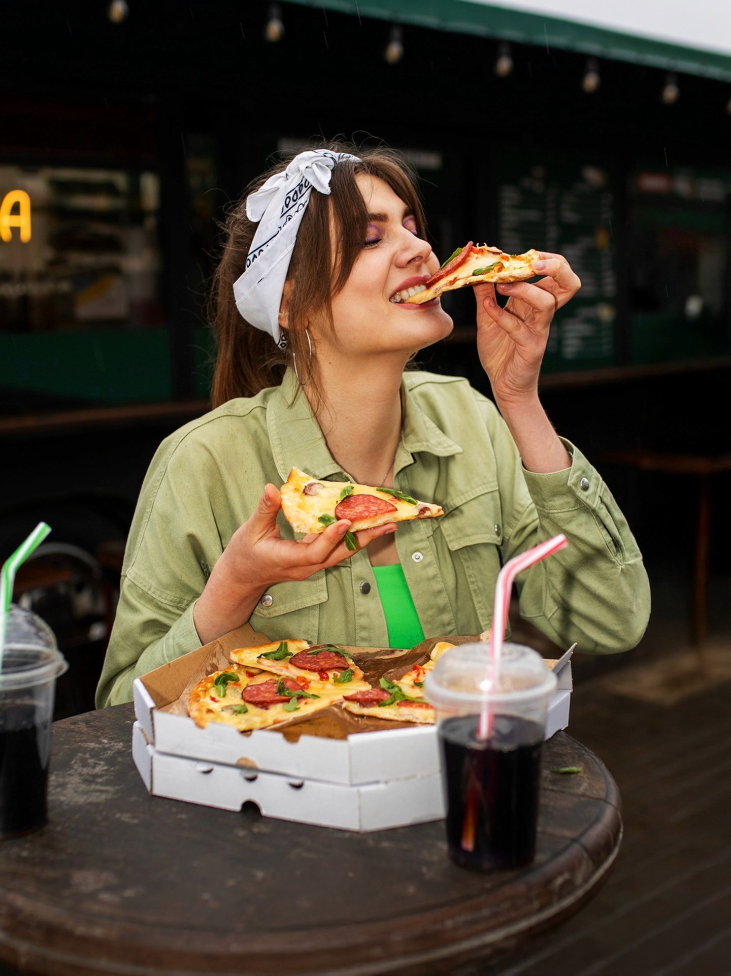 A young woman wearing a white bandana and green jacket happily eating a slice of pizza at an outdoor table, with an open pizza box and soda cups in front of her.