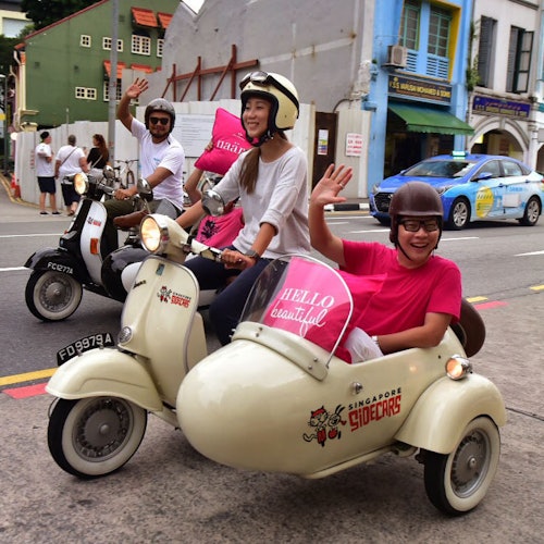 People riding scooters with sidecars, waving and smiling on a street with colorful buildings and other vehicles in the background.