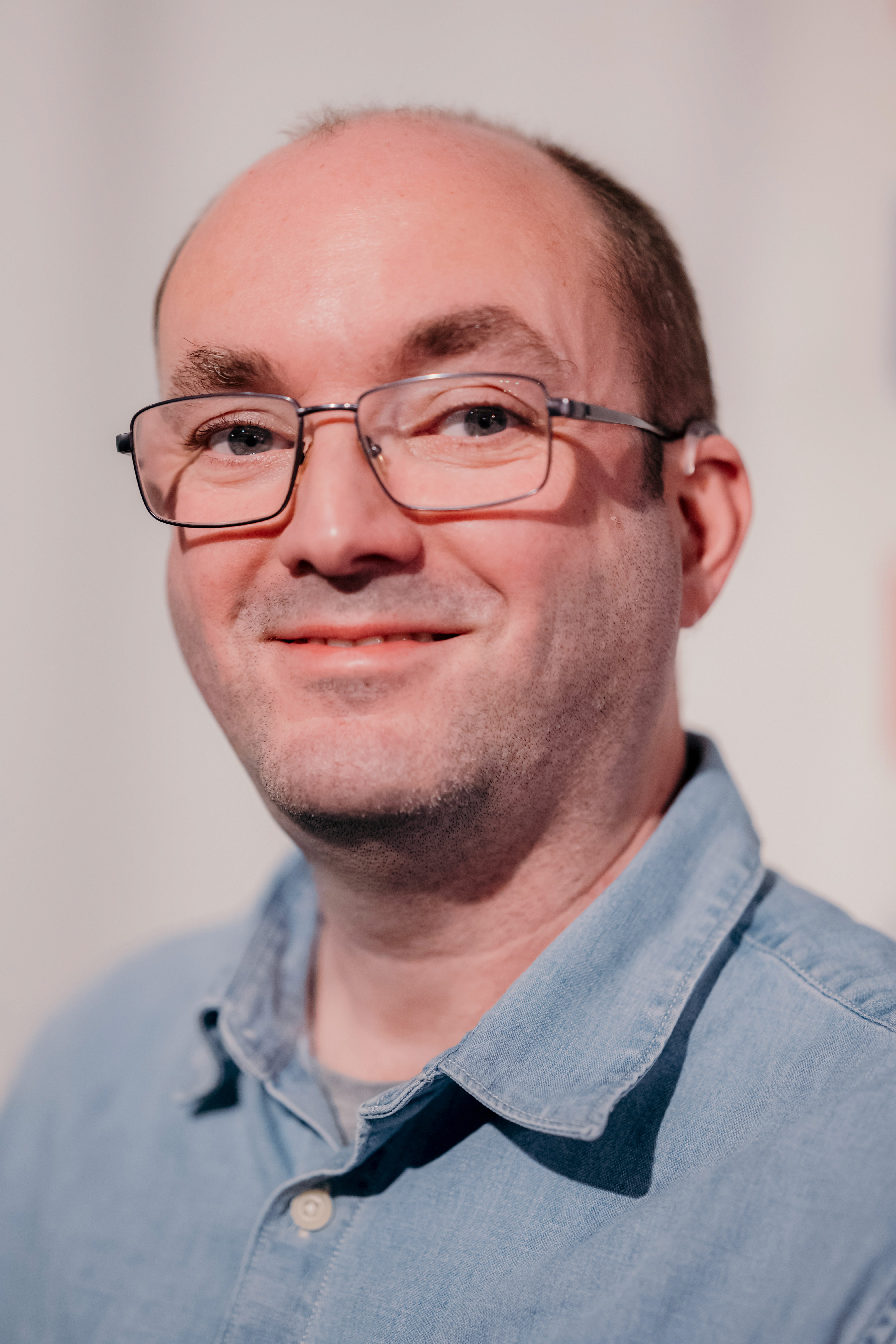 A man with short hair and glasses wearing a light blue polo shirt smiles at the camera in front of a white background