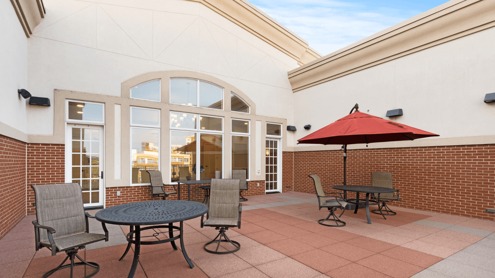 Spacious outdoor patio at The Gateway Village apartments for rent, featuring tiled flooring, multiple seating areas with tables and chairs, and a large red umbrella for shade.