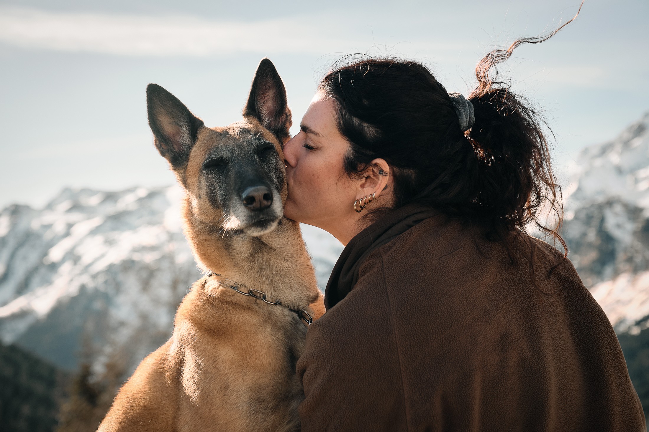A woman kisses her dog while enjoying a scenic mountain view with snow-capped peaks in the background.