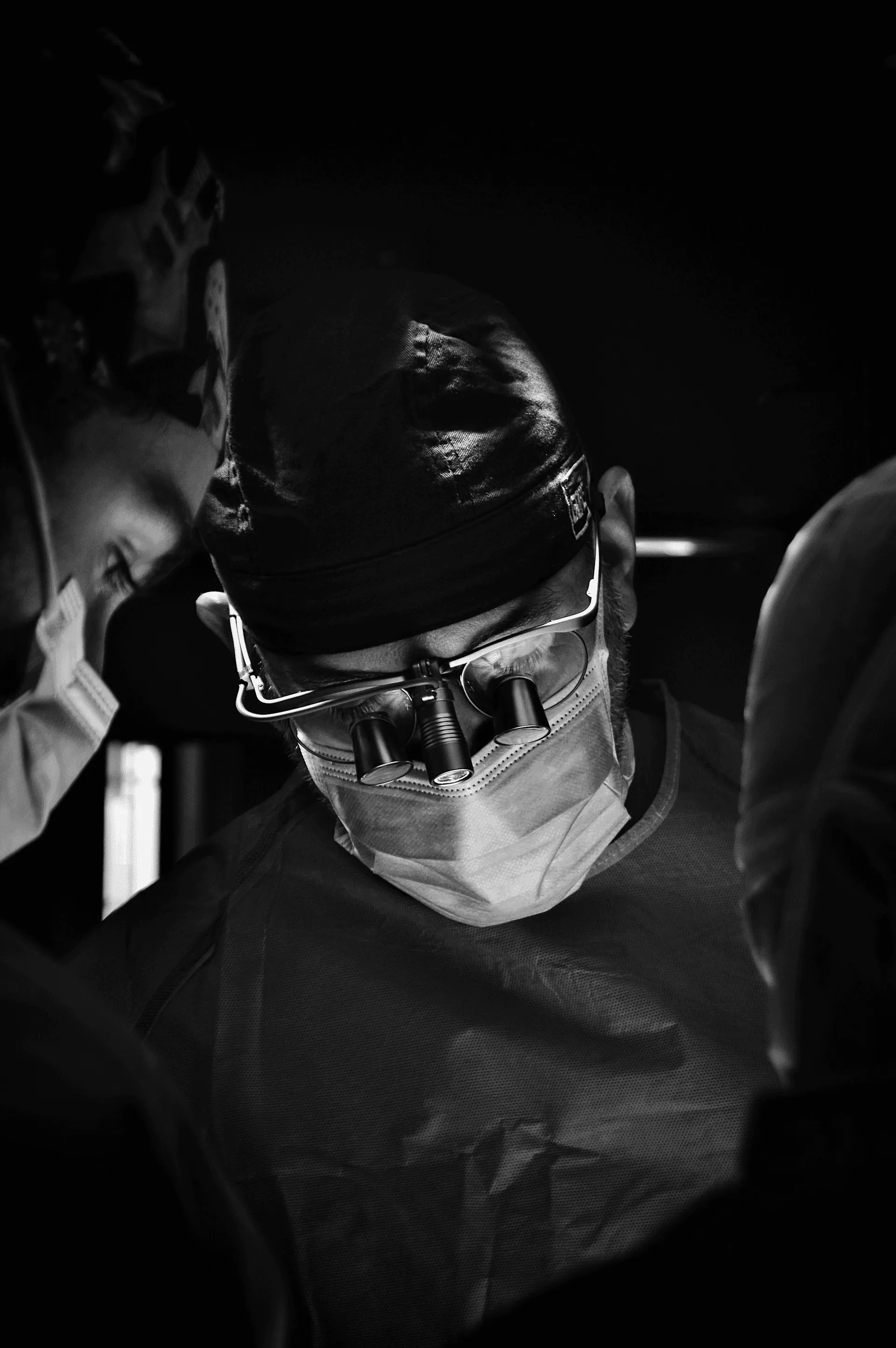 Black and white photo of a surgeon in a mask, cap, and magnifying loupes, looking down with intense focus