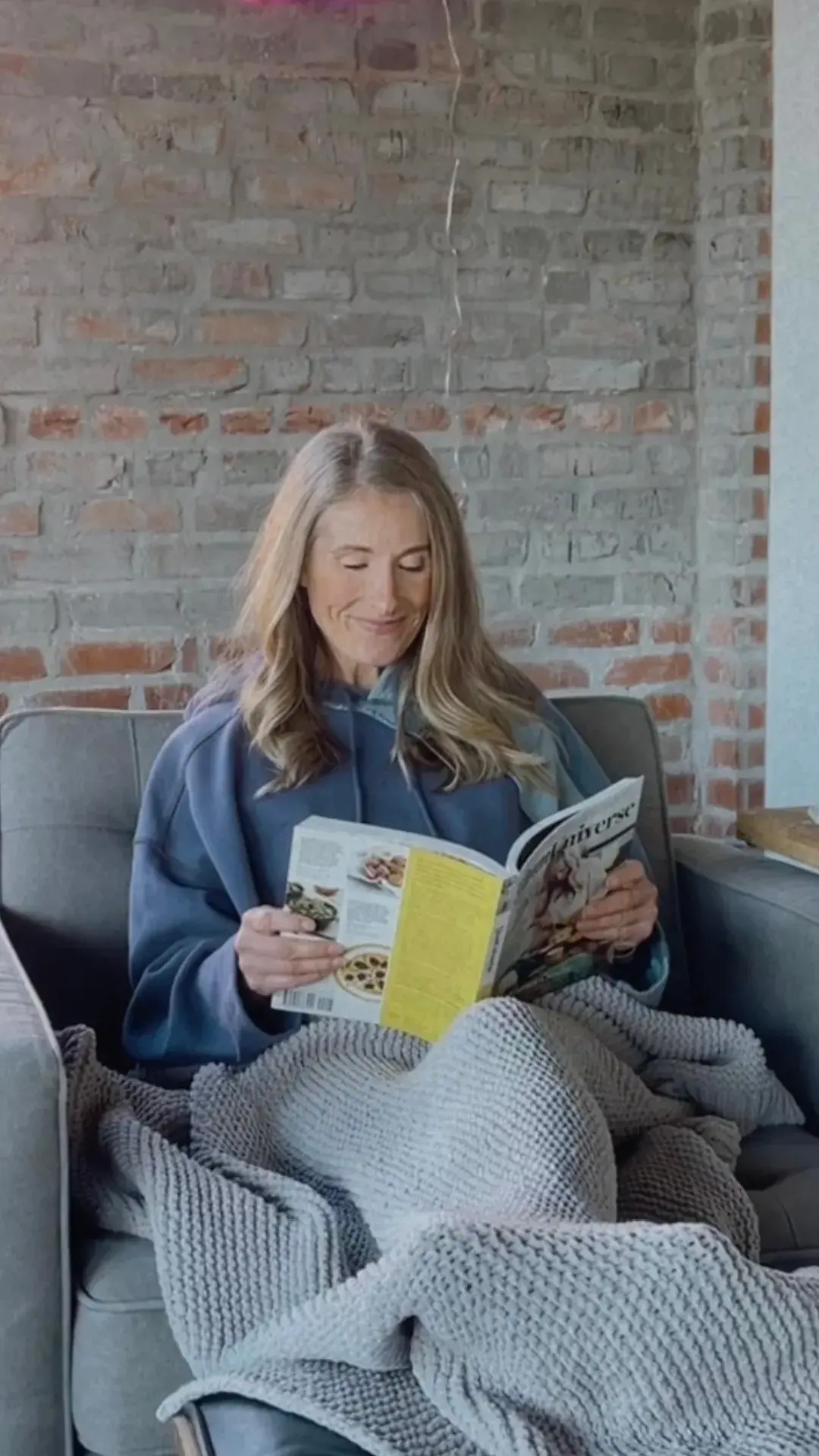 Heather Hausman with long hair is sitting comfortably on a gray sofa, wrapped in a textured gray blanket, reading a cooking magazine; a brick wall serves as a rustic backdrop.