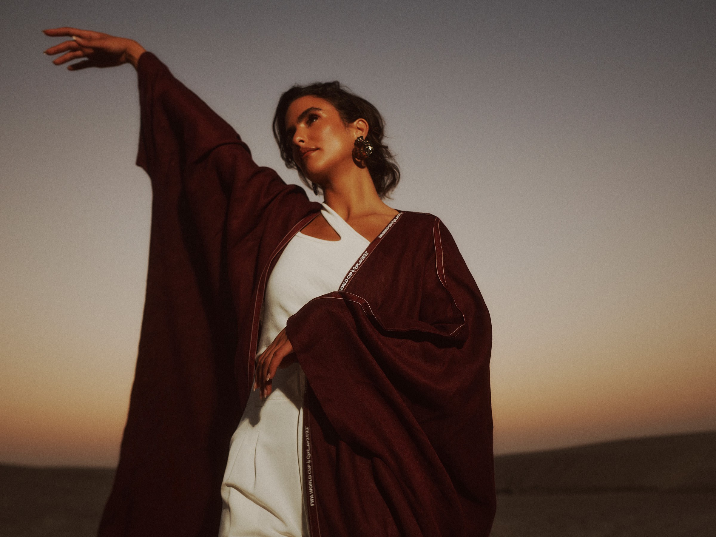 A model in a deep burgundy abaya over white, one arm raised against a desert sunset.