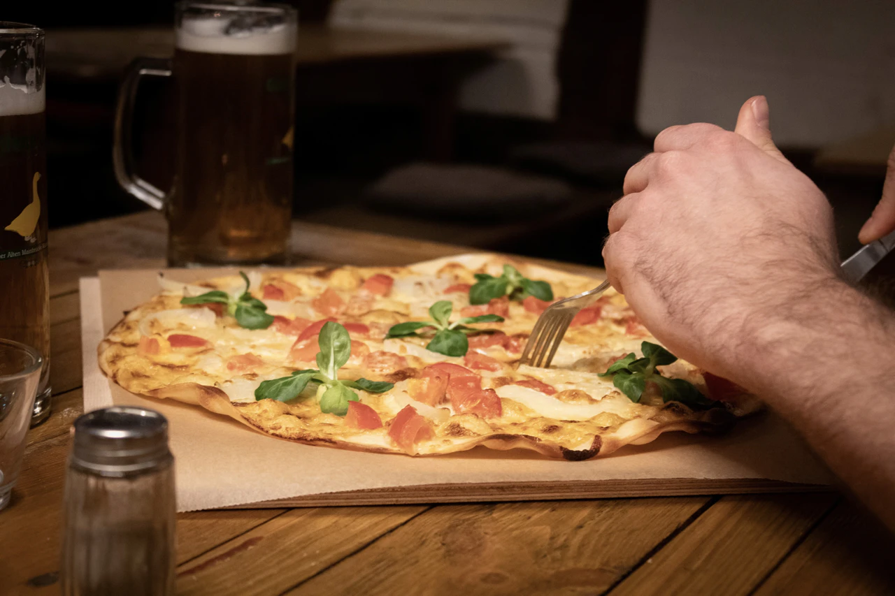 Flammkuchen mit Tomaten, Zwiebeln und Basilikum, serviert auf einem Holztisch; im Hintergrund zwei Biergläser.