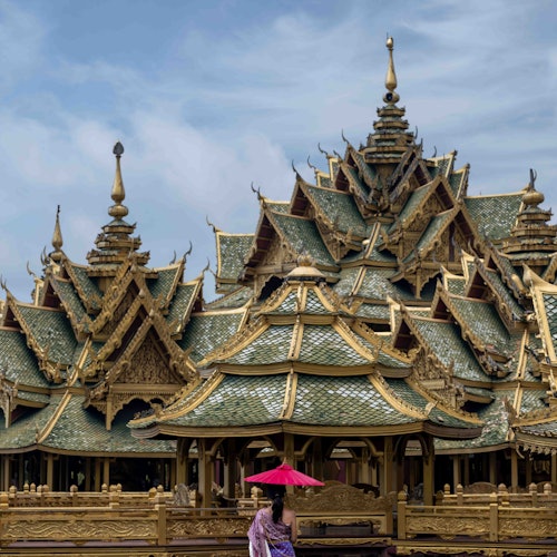 在多云的天空下，一位撑着红色雨伞的人站在一个细致、华丽并具有金色和绿色屋顶的寺庙建筑群前。