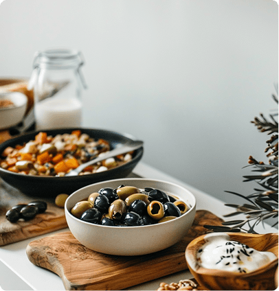 A close-up of a rustic food spread featuring a small bowl filled with mixed green and black olives on a wooden board, with a bowl of yogurt or dip and a salad in the background.